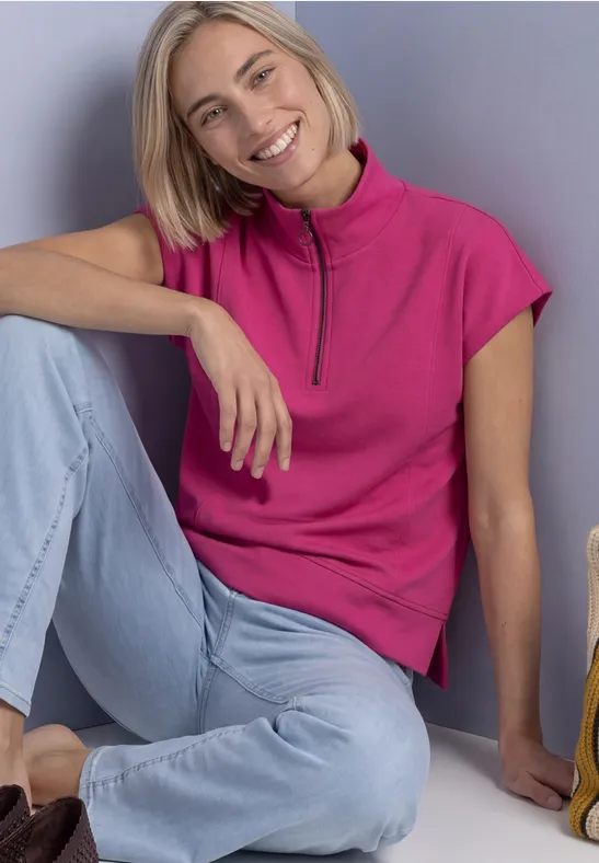 Eine lächelnde Frau sitzt auf dem Boden, lehnt an einer Wand, trägt pinkes Oberteil und helle Jeans.