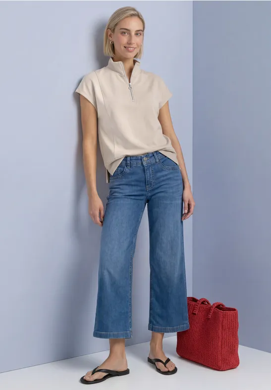Frau in beigem Kurzarmshirt und weiten Jeans mit Sandalen lehnt an hellblauer Wand, rote Tasche am Boden.