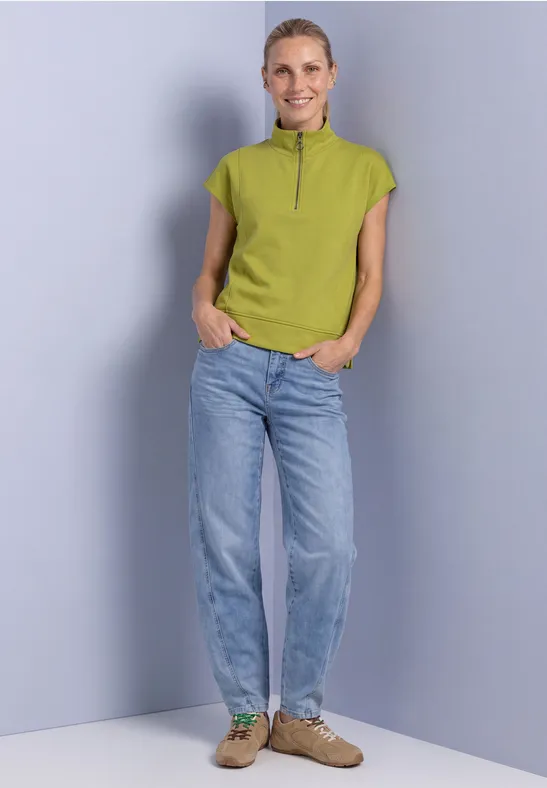 Eine Frau in hellblauer Jeans, grünem kurzärmeligem Shirt mit Reißverschluss und braunen Schuhen vor grauem Hintergrund.