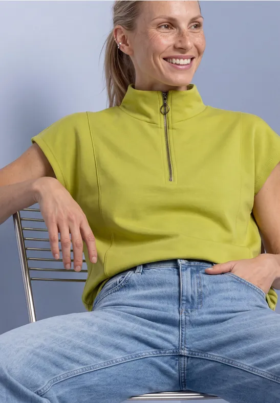 Eine lächelnde Frau in grünem Top und Jeans sitzt lässig auf einem Stuhl.