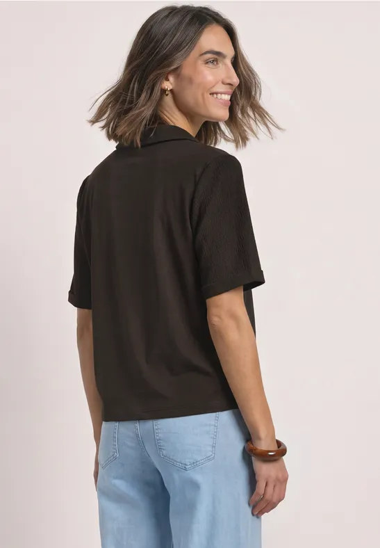 Frau mit kurzen braunen Haaren trägt schwarzes Kurzarm-Poloshirt und helle Jeans, von hinten sichtbar.