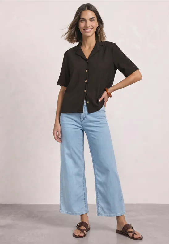 Frau lächelt in schwarzer Kurzarmbluse, hellblauen, weiten, knöchellangen Jeans und schwarzen Sandalen.