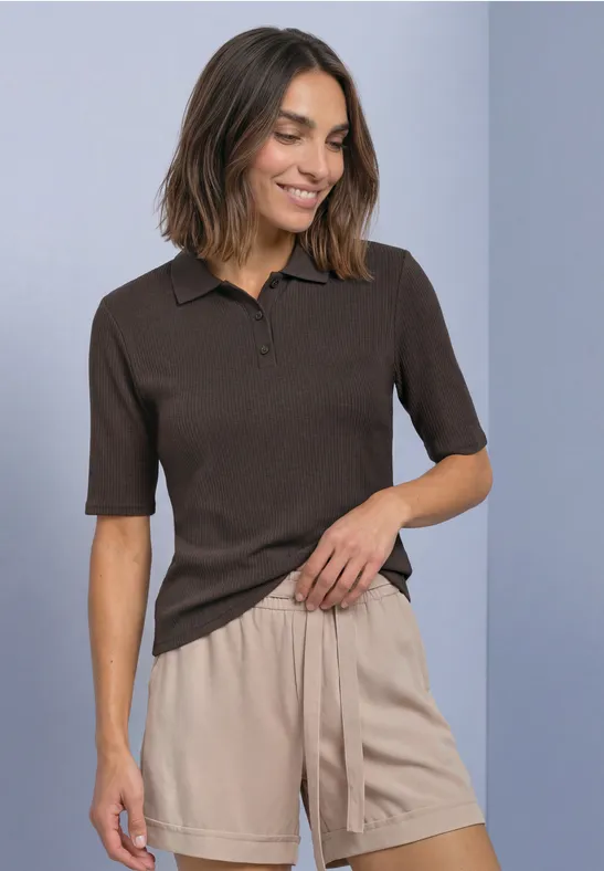 Frau im braunen Kurzarm-Poloshirt und beigefarbenen Shorts, lächelnd, eine Hand in der Tasche.