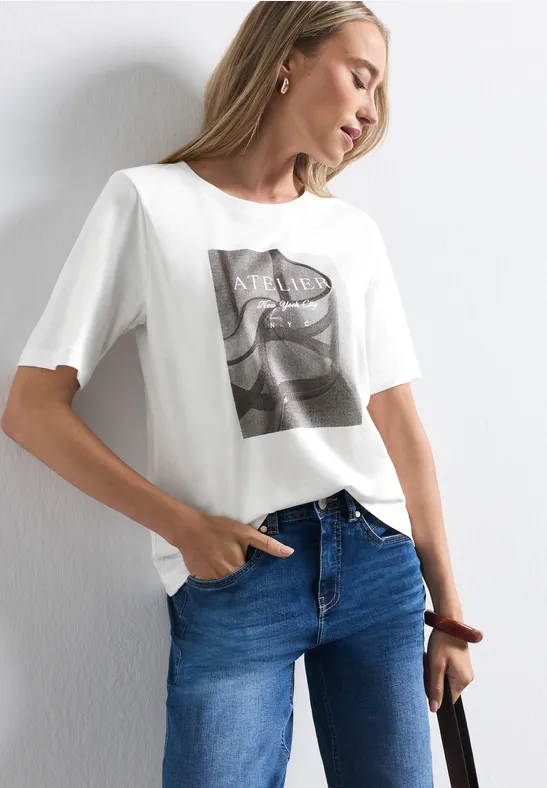 Frau in weißem T-Shirt mit Fotoprint, blauen Jeans und Tasche, vor einer weißen Wand posierend.