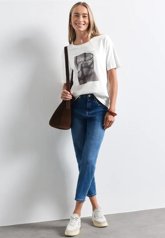 Eine junge Frau in Jeans, T-Shirt und Sneakers mit brauner Tasche lächelt vor weißem Hintergrund.
