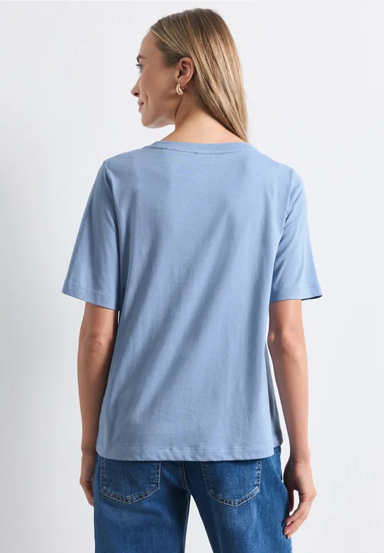 Eine Frau steht mit dem Rücken zur Kamera, sie trägt ein hellblaues T-Shirt und Jeans.
