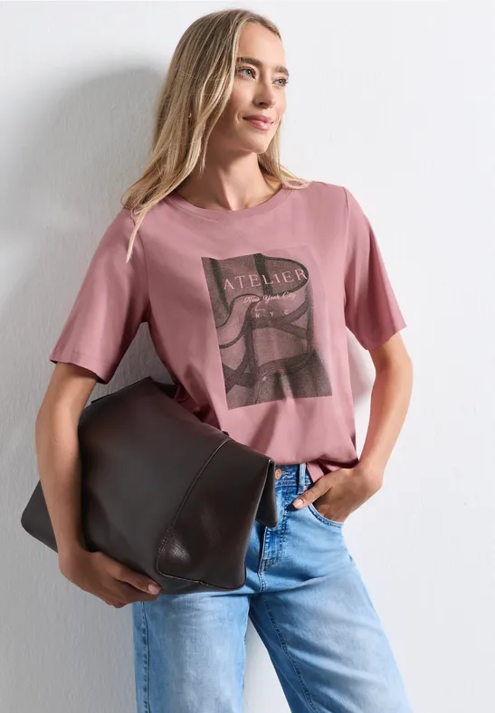 Frau in rosafarbenem T-Shirt mit Print, blauen Jeans und großer brauner Tasche vor weißem Hintergrund.