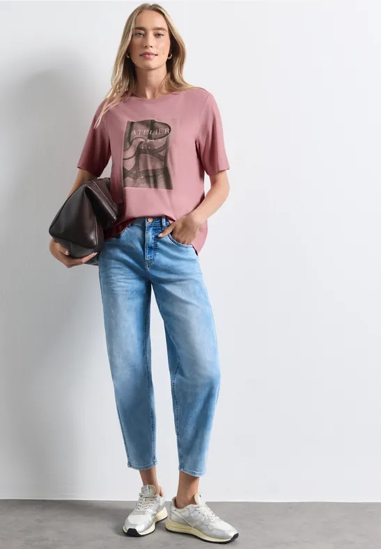 Frau trägt rosa T-Shirt mit Print, helle Jeans, Sneaker und hält eine schwarze Tasche.