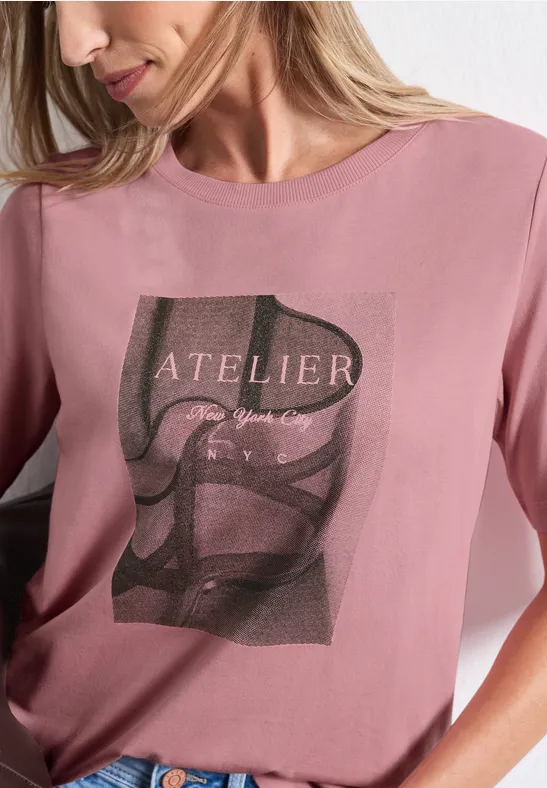 Frau in rosafarbenem T-Shirt mit Schriftzug 'ATELIER' und grafischem Schwarz-Weiß-Motiv.
