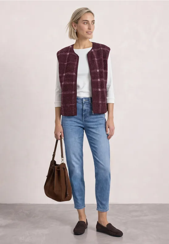 Frau trägt weiße Bluse, burgunderfarben gesteppte Weste, Jeans, schwarze Loafers und braune Tasche.
