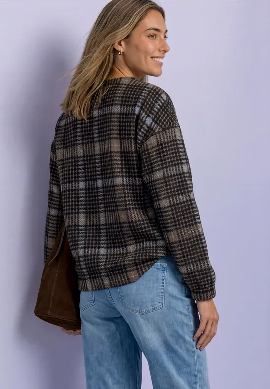 Femme en pull à carreaux et jean sourit en regardant par-dessus son épaule devant un mur clair.