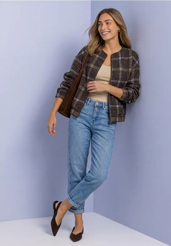 Femme en jean, top beige et veste à carreaux, souriante devant un fond clair.