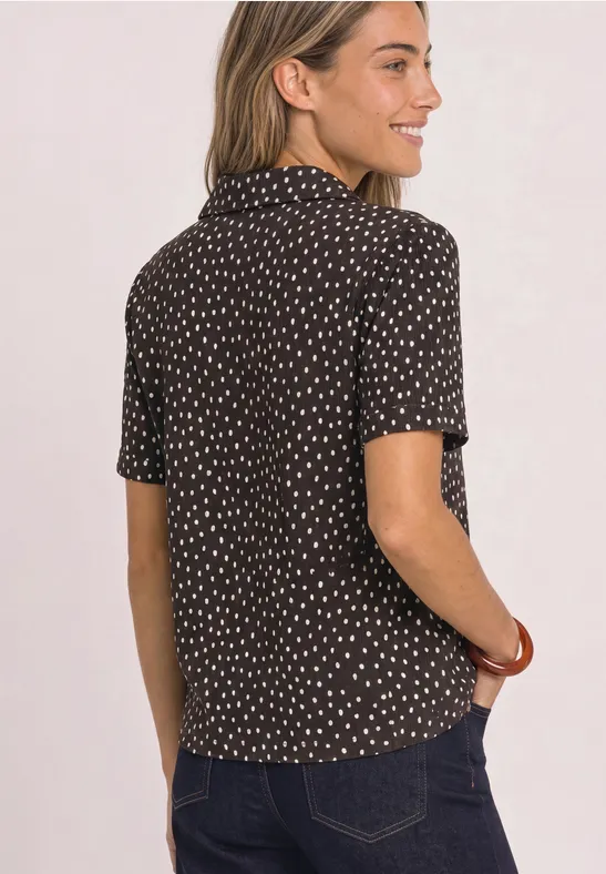 Frau von hinten in schwarzer Bluse mit weißen Punkten und Jeans, die lächelnd zur Seite blickt.