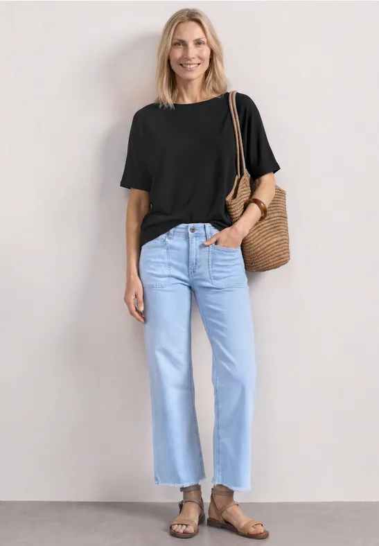 Frau in schwarzem T-Shirt, hellblauen Jeans und Sandalen trägt eine geflochtene Strohtasche vor weißem Hintergrund.