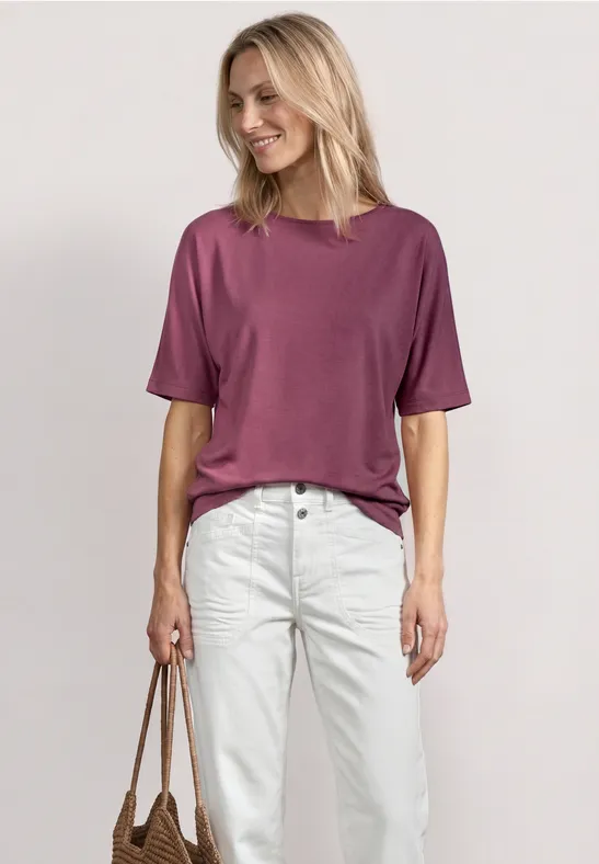 Eine Frau steht vor neutralem Hintergrund, trägt ein violettes T-Shirt, weiße Jeans und hält eine braune Tasche.