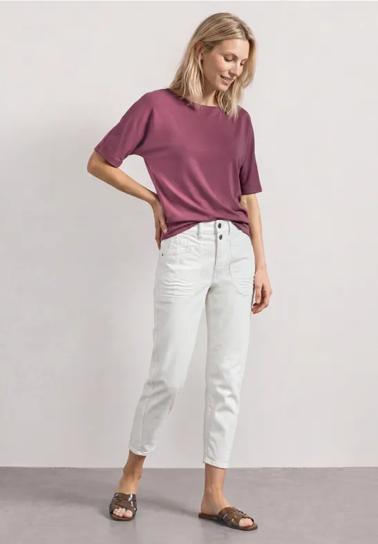 Frau in lässiger Pose mit lila T-Shirt, hellen Jeans und Sandalen vor neutraler Wand.