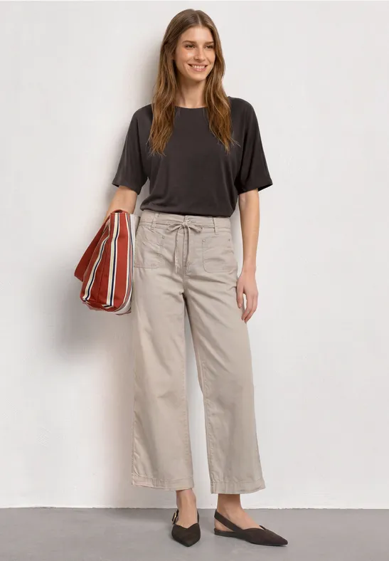 Vrouw in zwart T-shirt, beige wijde broek, zwarte ballerina’s en rood gestreepte shopper.