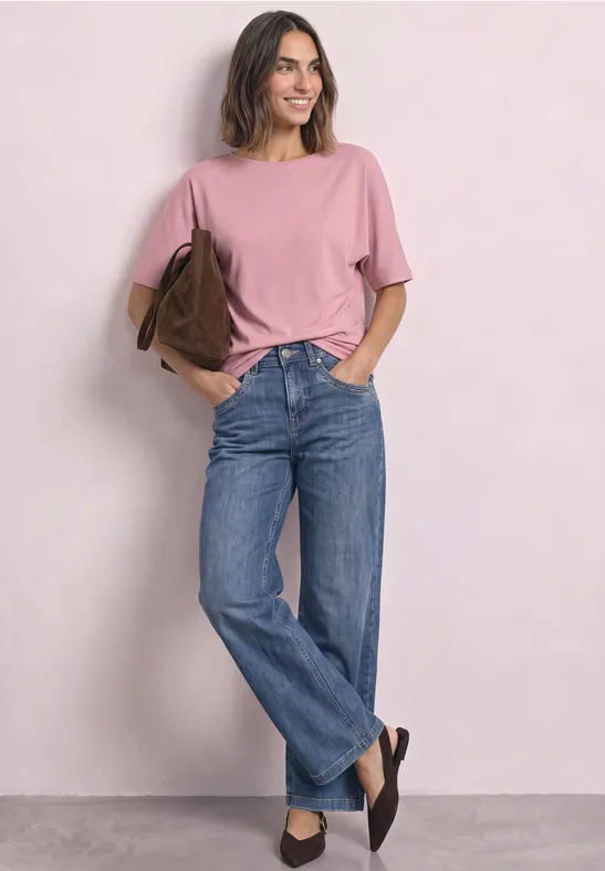 Kobieta w różowym T-shircie, niebieskich jeansach i czarnych butach, uśmiechnięta z brązową torbą na ramieniu.