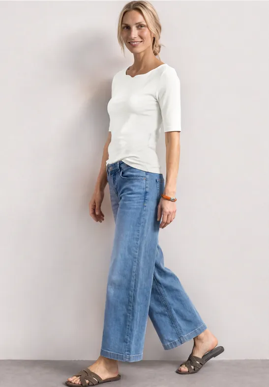 Vrouw in wit 3/4-shirt, wijde blauwe jeans en sandalen tegen een neutrale achtergrond.