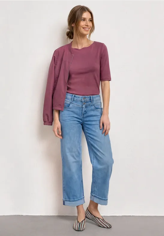 Une femme porte un top et une veste violets sur l'épaule, un jean ample et des slingbacks rayés.
