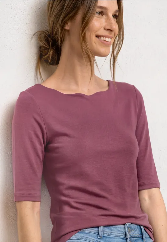 Une femme porte un t-shirt rose à manches mi-longues devant un fond blanc.