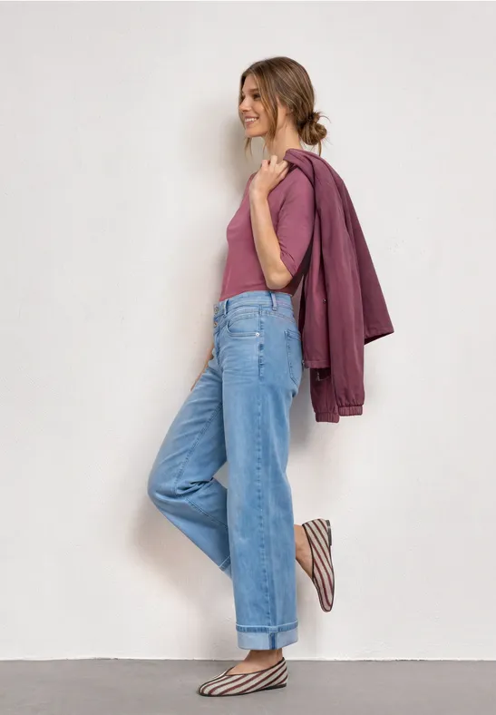 Femme souriante appuyée contre un mur blanc, porte un top rose, une veste violette, jean large bleu et slip-ons rayés.