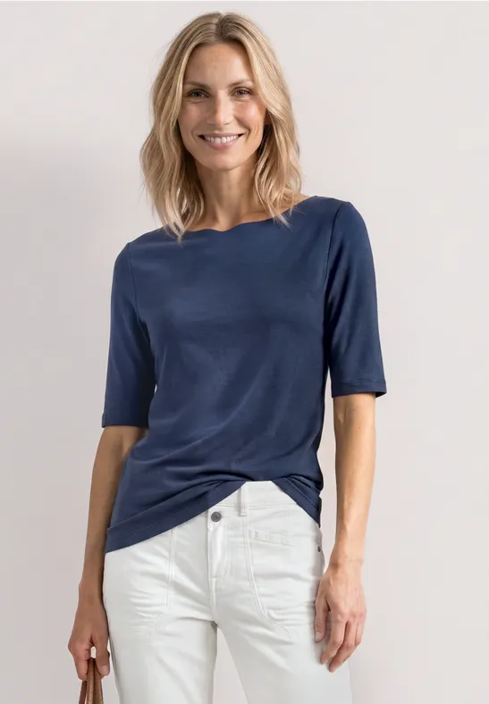Une femme porte un t-shirt bleu marine et un pantalon blanc devant un fond clair.