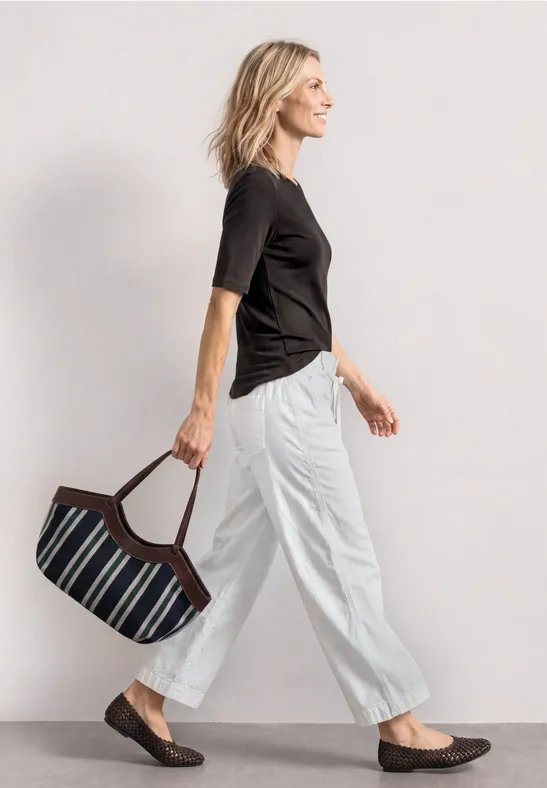 Vue de profil d’une femme en t-shirt noir et pantalon blanc, portant un sac à main rayé.