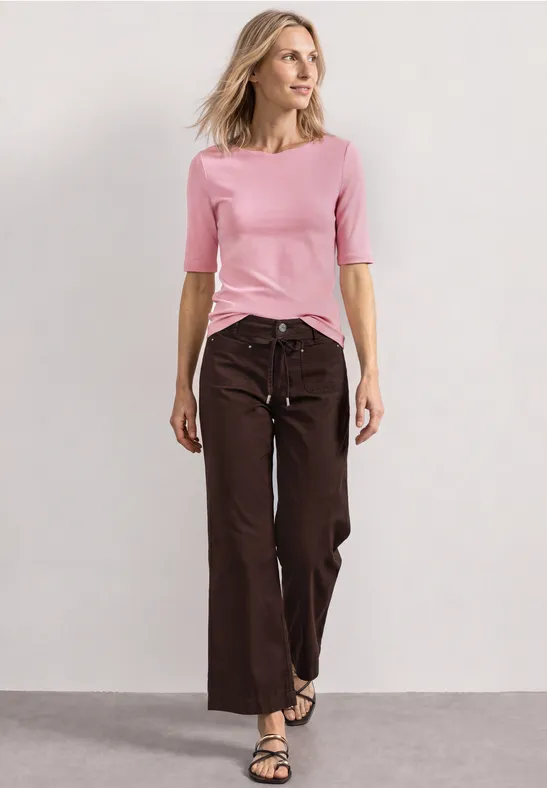 Vrouw in roze top en brede bruine broek staat voor een neutrale achtergrond.