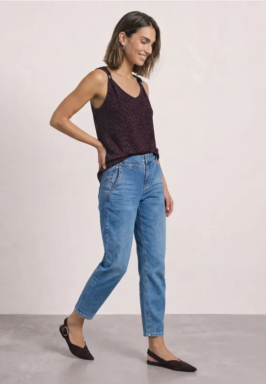 Eine junge Frau lächelt in schwarzem Top, blauen High-Waist-Jeans und schwarzen Ballerinas.