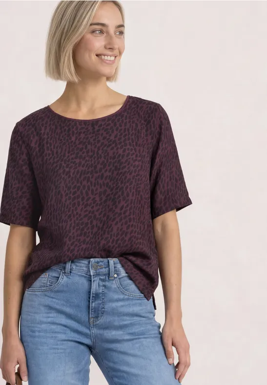Kobieta w fioletowym, wzorzystym T-shircie i dżinsach, uśmiechnięta na jasnym tle.