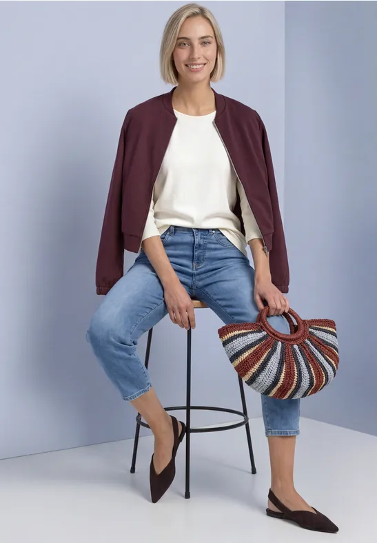 Frau in weißem Shirt, Jeans und burgunderrotem Jäckchen sitzt auf einem Hocker und hält eine bunte Korbtasche.