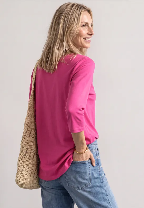 Frau in pinkem 3/4-Arm-Shirt, Jeans und gehäkelter Tasche, seitlich stehend und zurückblickend.