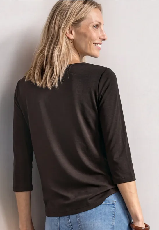 Frau mit blonden Haaren trägt braunes 3/4-Arm-Shirt und Jeans, lächelnd seitlich