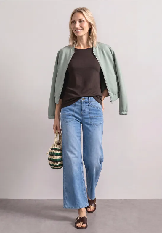 Dame in lockerer Jeans, schwarzem Top und grüner Jacke, trägt Korbtasche und Sandalen vor hellem Hintergrund.