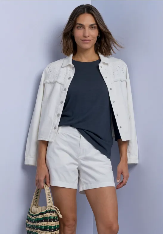 Frau mit weißer Jeansjacke, dunkelblauem T-Shirt, weißen Shorts und geflochtenem Korb vor hellgrauem Hintergrund.