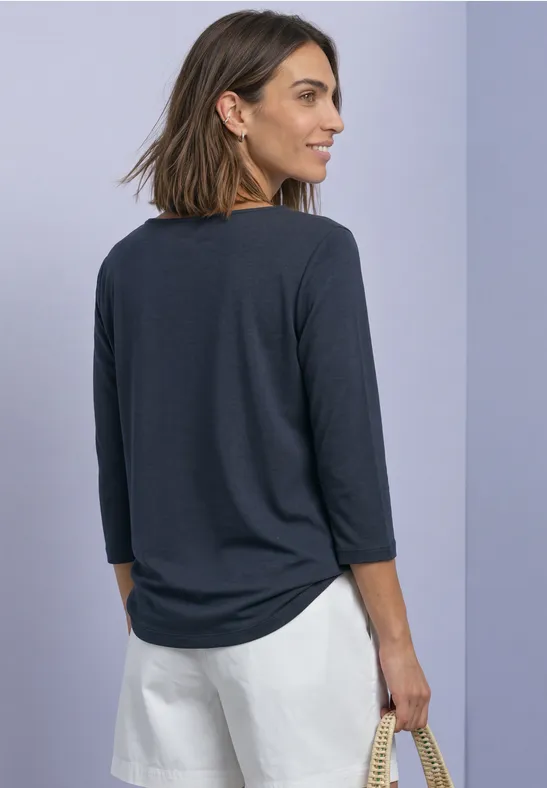 Frau in dunkelgrauem 3/4-Arm-Shirt und weißen Shorts, seitlich gewandt mit Korbtasche in Hand vor lila Hintergrund.