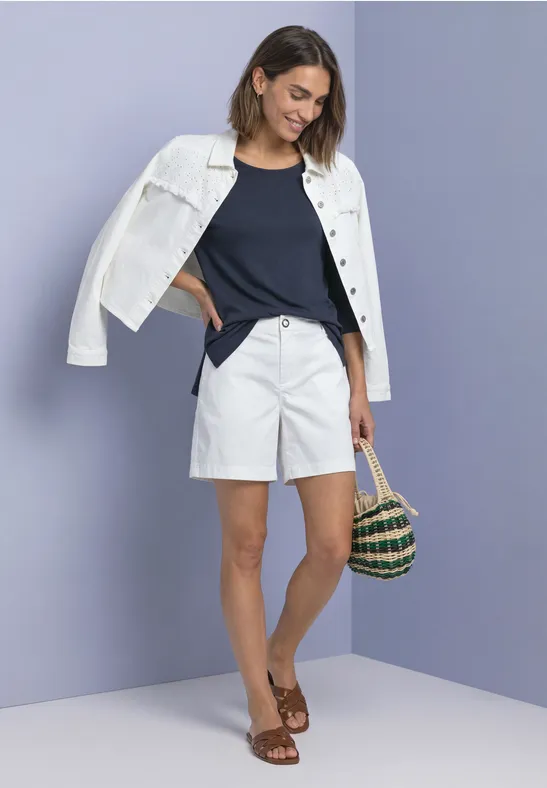 Frau mit weißer Jacke auf den Schultern, dunkelblauem Top, weißen Shorts, braunen Sandalen und Korbtasche vor lila Wand.
