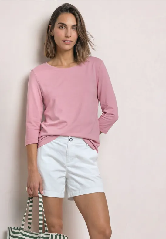Frau in rosafarbenem 3/4-Arm-Shirt, hellen Shorts und gestreifter Tasche vor einfarbigem Hintergrund.