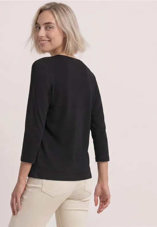 Frau von hinten, lächelnd über die Schulter blickend, trägt schwarzen 3/4-Arm-Pullover und helle Hose.