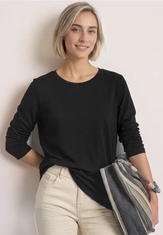 Frau mit kurzem blonden Haar trägt schwarzen Langarmshirt, beigefarbene Hose und hält eine gestreifte Tasche.