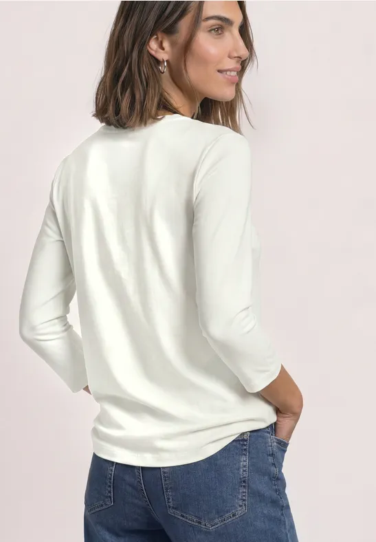Frau mit schulterlangem Haar, trägt weißen 3/4-Arm-Pullover und Jeans, steht seitlich und schaut.