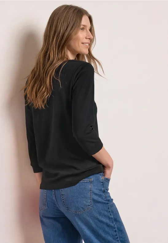 Eine Frau von hinten in schwarzem Oberteil und blauen Jeans, die entspannt an einer Wand steht.
