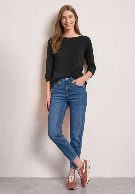 Eine Frau in schwarzem Langarmshirt, blauen Jeans und Sneakers steht lässig vor einer hellen Wand.