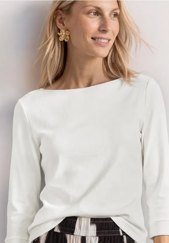 Frau trägt weiße 3/4-Ärmel-Bluse mit U-Boot-Ausschnitt, gemusterten Rock und Blumenohrring.