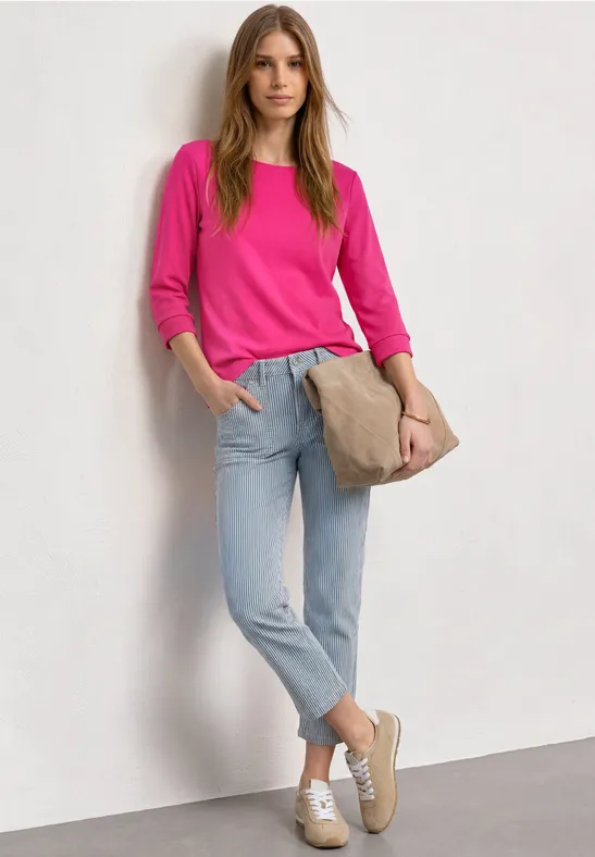 Junge Frau lehnt an weißer Wand, trägt pinkes Shirt, helle Jeans, weiße Sneaker und eine braune Umhängetasche.
