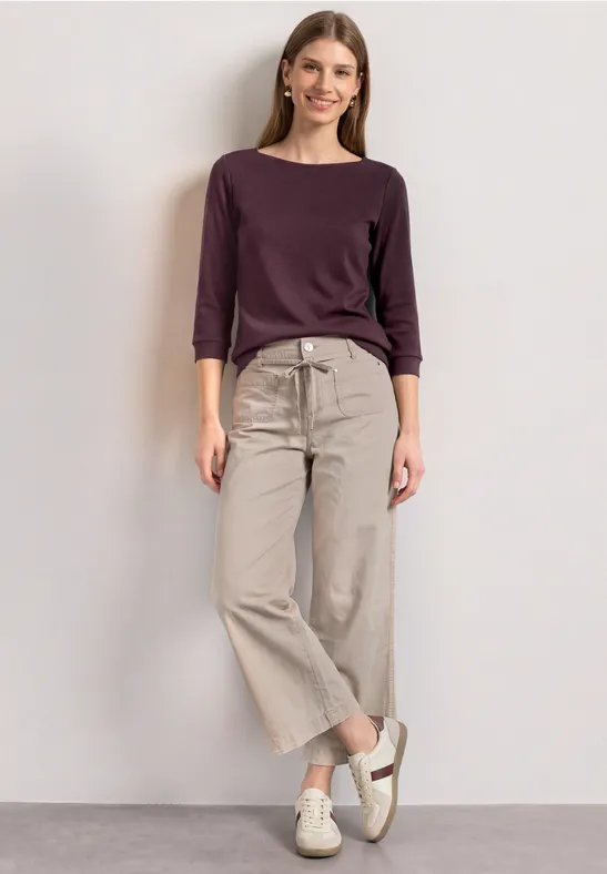 Frau in lila 3/4-Arm-Shirt und beiger weiter Hose mit weißen Schuhen vor hellem Hintergrund.