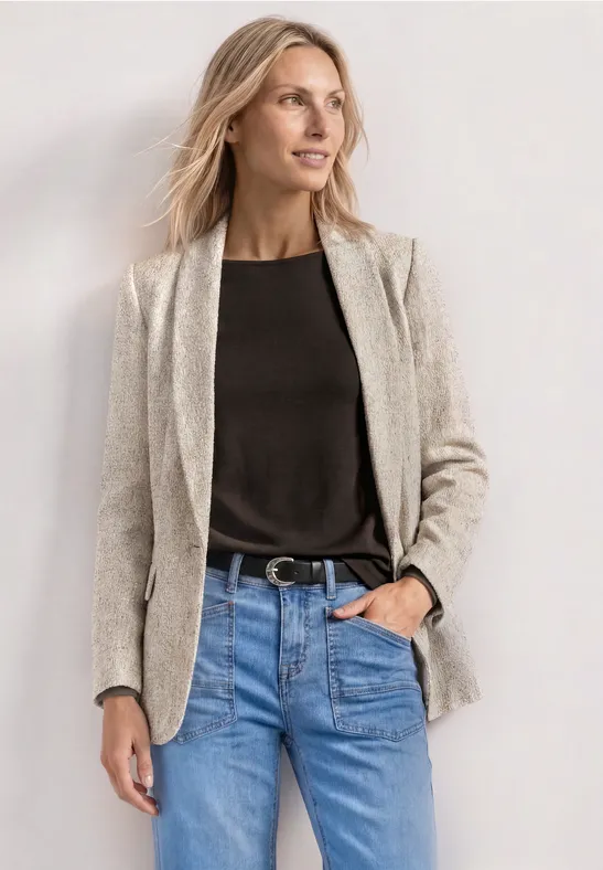 Frau in beigem Blazer, schwarzem Shirt, blauen Jeans und Gürtel posiert vor weißem Hintergrund.