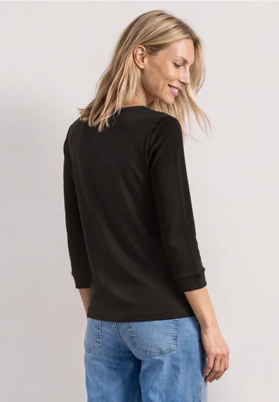Eine Frau steht von hinten, trägt schwarzen 3/4-Arm-Pullover und blaue Jeans.