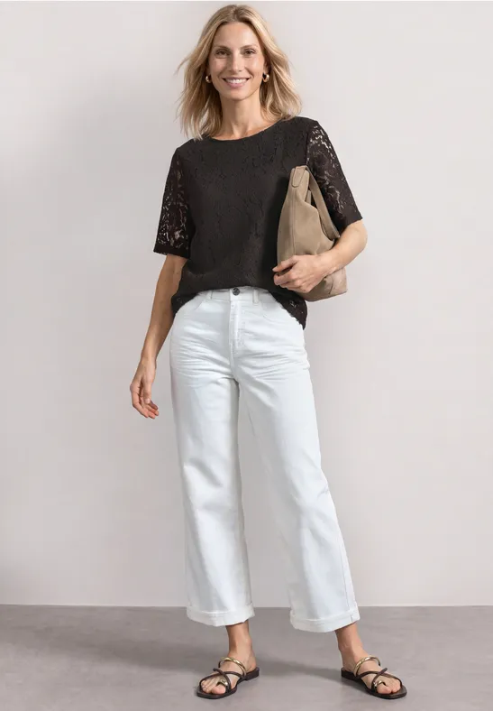 Frau in schwarzer Spitzenbluse, weißer Jeans, schwarzen Sandalen, lächelnd mit beiger Clutch vor neutralem Hintergrund.